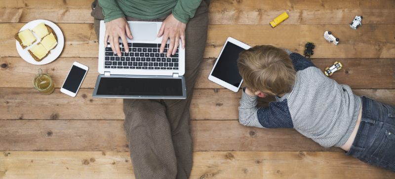 Lap of adult with laptop; small child laying on floor with little cars & iPad mini